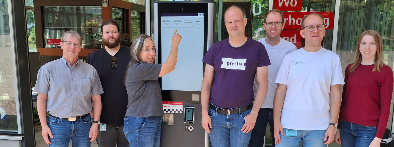 Eine Gruppe Menschen steht links und rechts von einem Ticketautomaten und guckt in die Kamera, eine Frau mit grauen Haaren tippt auf den Bildschirm des Automaten